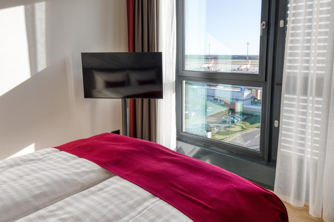 Intercityhotel Berlin Airport Ber Terminal 1 & 2 Schönefeld Exteriér fotografie The photo shows a modern hotel room with a large bed covered in white linens and a burgundy throw blanket. In the background, there is a large window that lets in natural light, offering a view of the outside. A flat-screen TV is mounted on the wall 