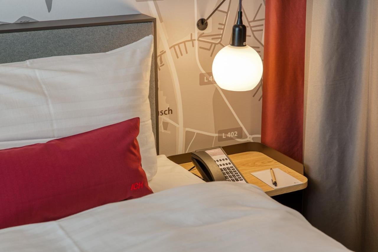 Intercityhotel Berlin Airport Ber Terminal 1 & 2 Schönefeld Exteriér fotografie The photo shows a cozy bedroom setup. There's a neatly made bed with white bedding and a red decorative pillow. Beside the bed, a small bedside table holds a telephone and a notebook or notepad, along with a pen. A round pendant light hangs above the