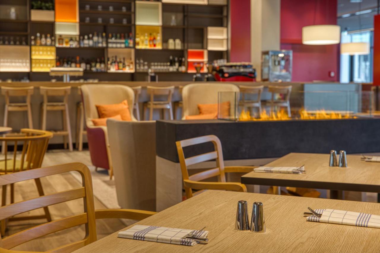 Intercityhotel Berlin Airport Ber Terminal 1 & 2 Schönefeld Exteriér fotografie The photo shows a modern restaurant or bar interior. In the foreground, there are wooden tables set with napkins and condiments. The seating includes wooden chairs and plush seating in various colors in the background. A stylish bar area is visible w
